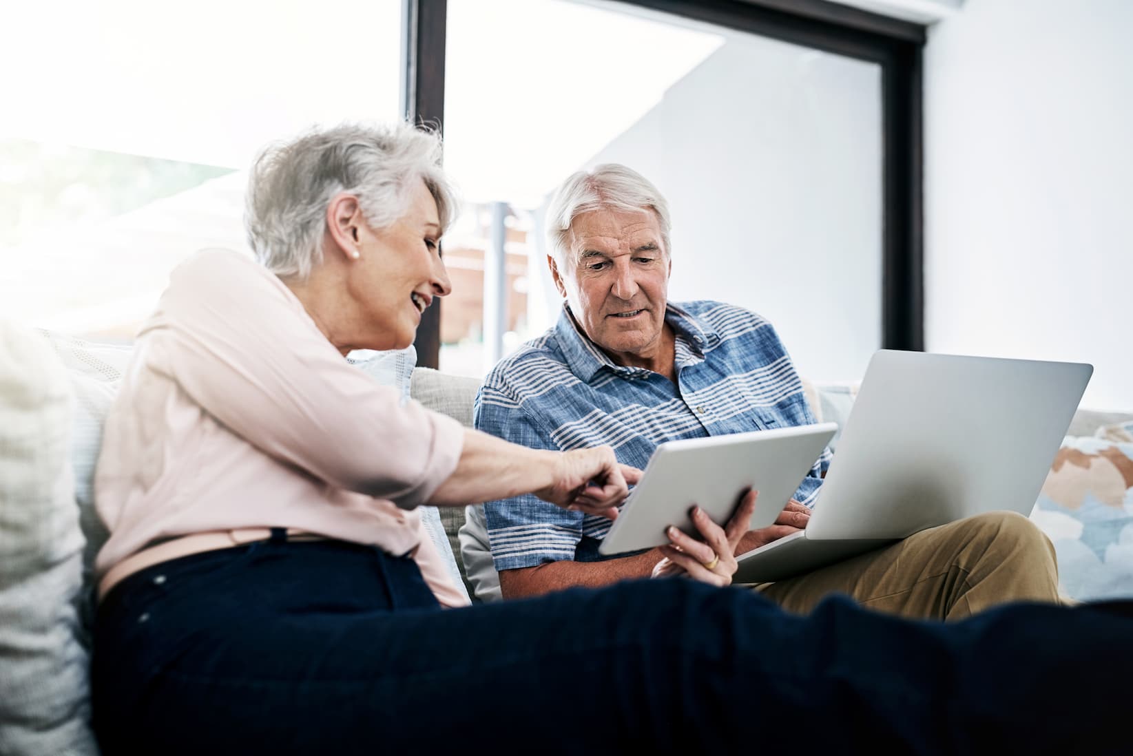 senior-couple-reading-online Senior man and woman sit on a couch and review information on a laptop and tablet screen together