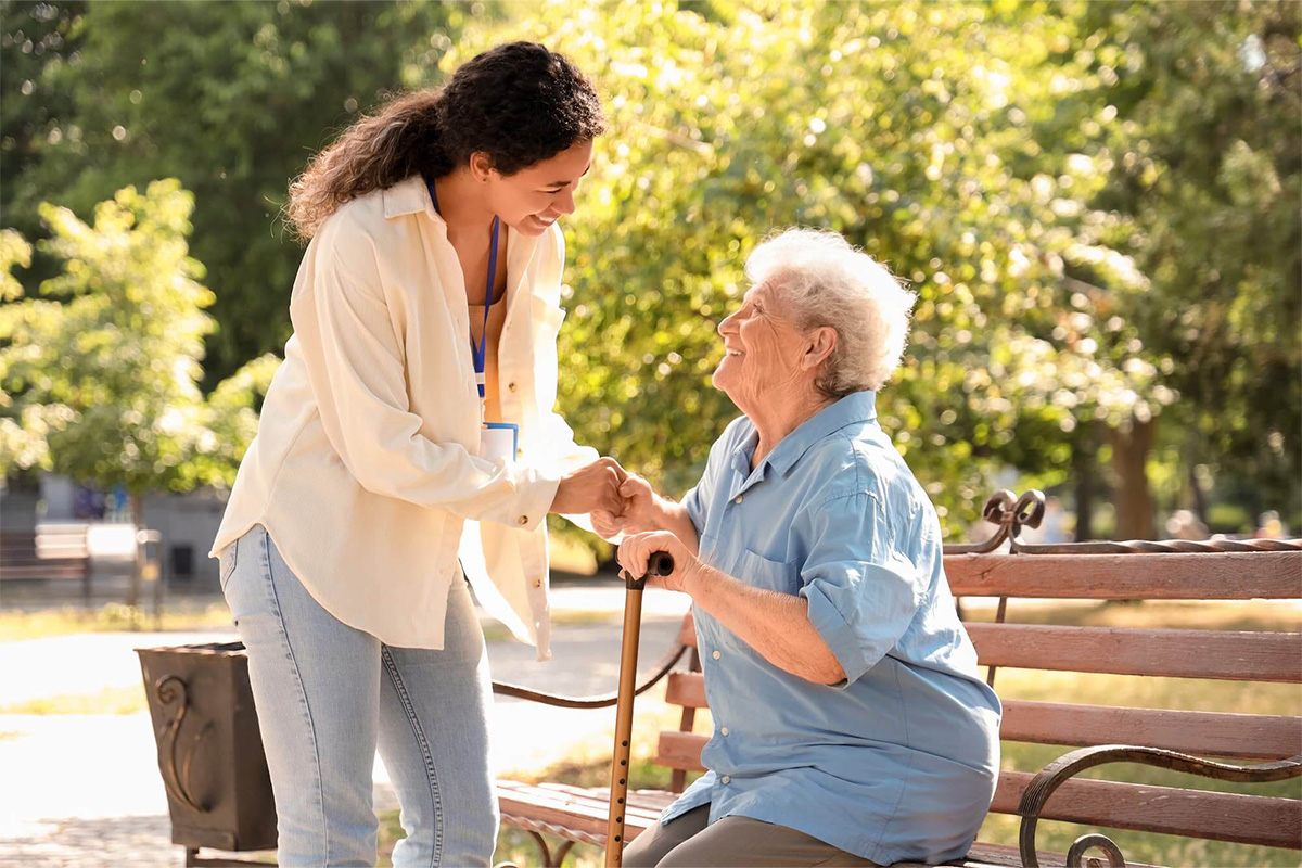 senior-woman-assisted-by-young-woman-outdoors Senior woman sits on an outdoor bench smiling up at younger female caretaker.